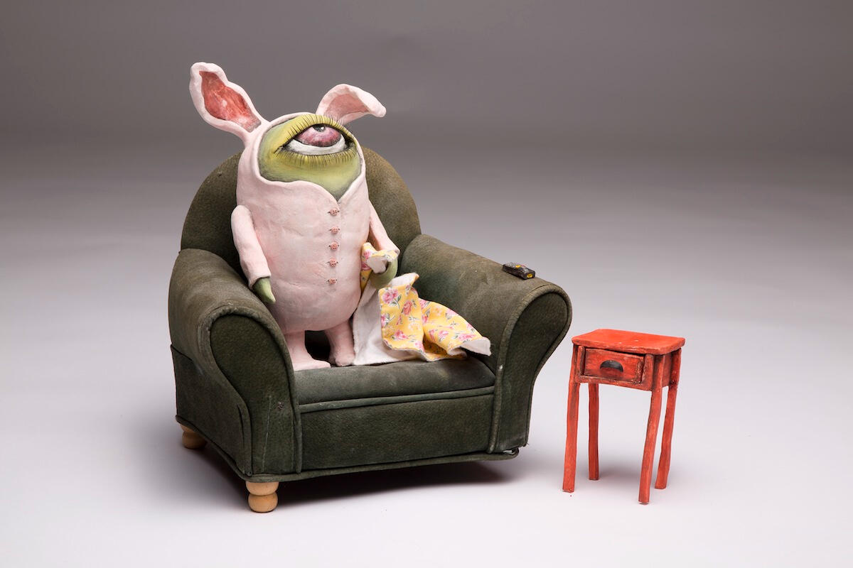 Sarah Alonzo Artist - Ceramics Ceramic green monster with one eye in a bunny suit. standing on a sofa chair with side table.